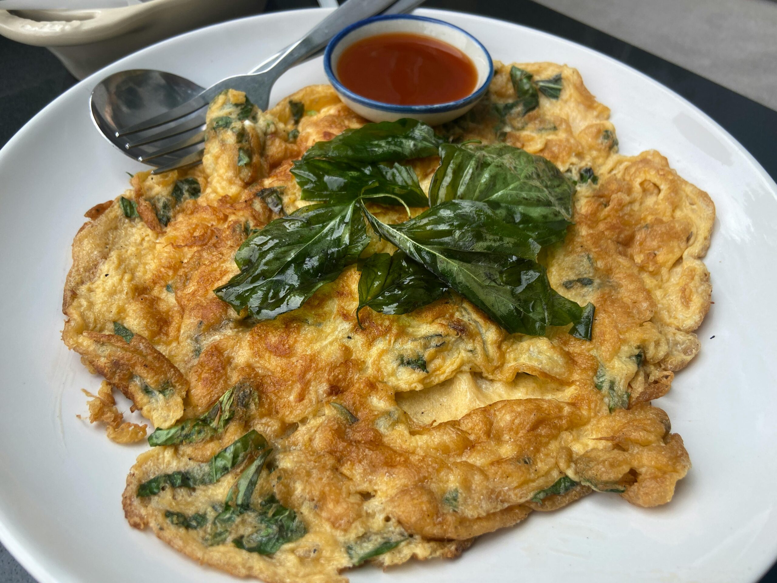 Our Menu Thai-style spinach omelette with basil leaves served with spicy sauce on a white plate.